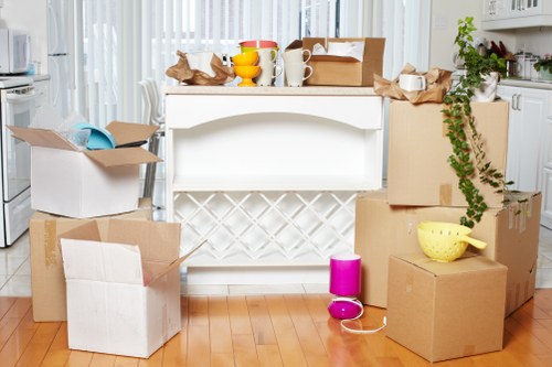 A kitchen scene featuring a white kitchen island with a lattice-style panel in the front, surrounded by multiple cardboard moving boxes of various sizes, some open with packing paper and miscellaneous household items visible. On top of the island are additional packing supplies, including a yellow bowl, a white mug, a small yellow container, and crumpled brown packing paper. To the right, a large cardboard box has a green vine-like plant draping over its edge, and a yellow plastic colander is placed on top. A pink and purple portable speaker and a white charging cable are on the floor among the boxes. The background reveals part of a modern kitchen with white cabinets, a window letting in natural light, and a glimpse of other household kitchen items, indicating a residential interior during a house move within the Swiss Cottage area. The scene is well-lit, reflecting an active packing process typical of house removals, with a clean and organized appearance suited for professional removals services by Swiss Cottage Man with Van.