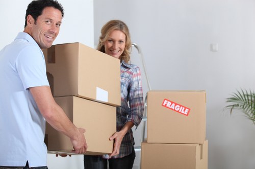 A man and a woman are indoors, participating in a house moving process; the man, dressed in a light blue shirt, is smiling and holding three cardboard boxes stacked in his hands, with the top box partially obscuring his face. The woman, wearing a plaid shirt, stands behind him, also smiling, with her hands on the boxes. Several more cardboard boxes are stacked to their right, some sealed and one bearing a red and white 'Fragile' sticker. The boxes range in size and are made of brown corrugated cardboard, with some showing white labels. The scene takes place in a bright, spacious interior with white walls and minimal decor, including a green potted plant visible on the far right. The environment suggests a residential setting, likely during a residential move or relocation activity, fitting the context of house removals services in the Swiss Cottage area, NW3, as offered by Swiss Cottage Man with Van. The overall lighting is natural, and the atmosphere is organized and professional, illustrating the process of packing and moving household items.
